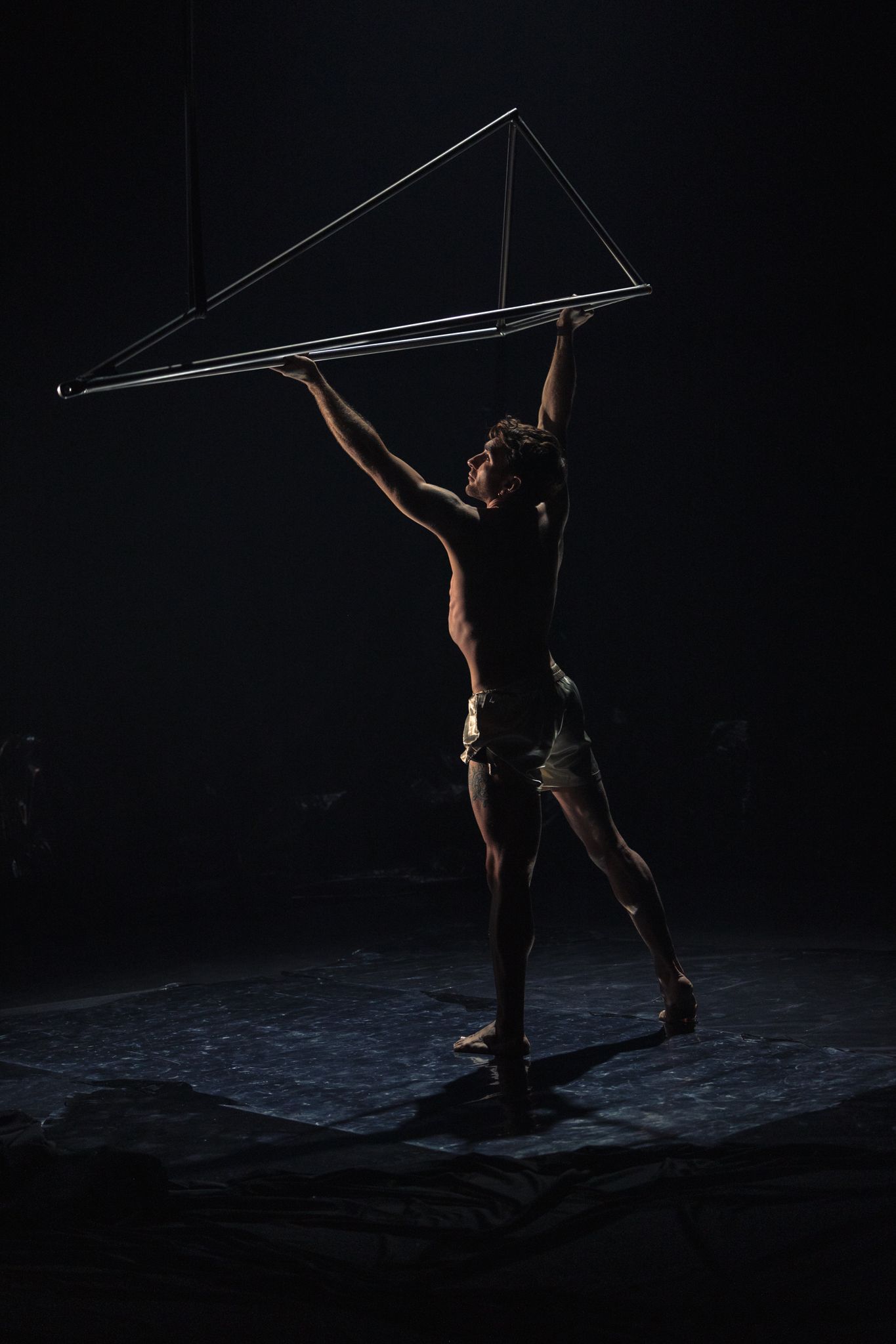A performer in dramatic lighting dances with a large geometric triangular light structure on a dark stage.