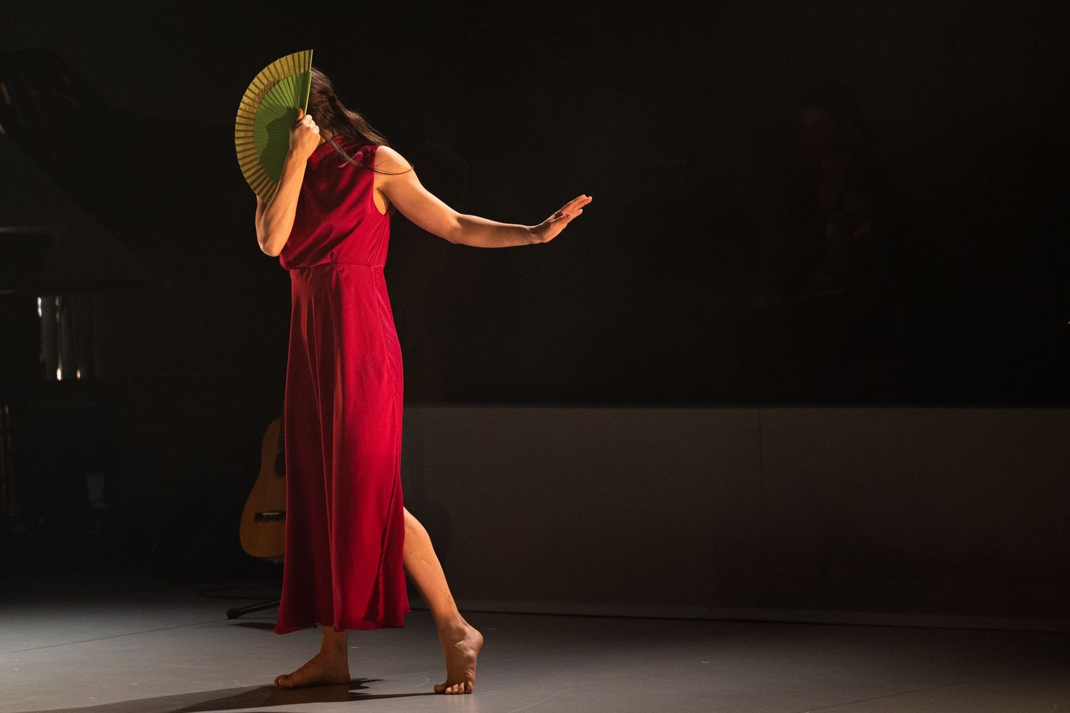 A dancer in a flowing red dress and hat performs an elegant pose on a dimly lit stage.