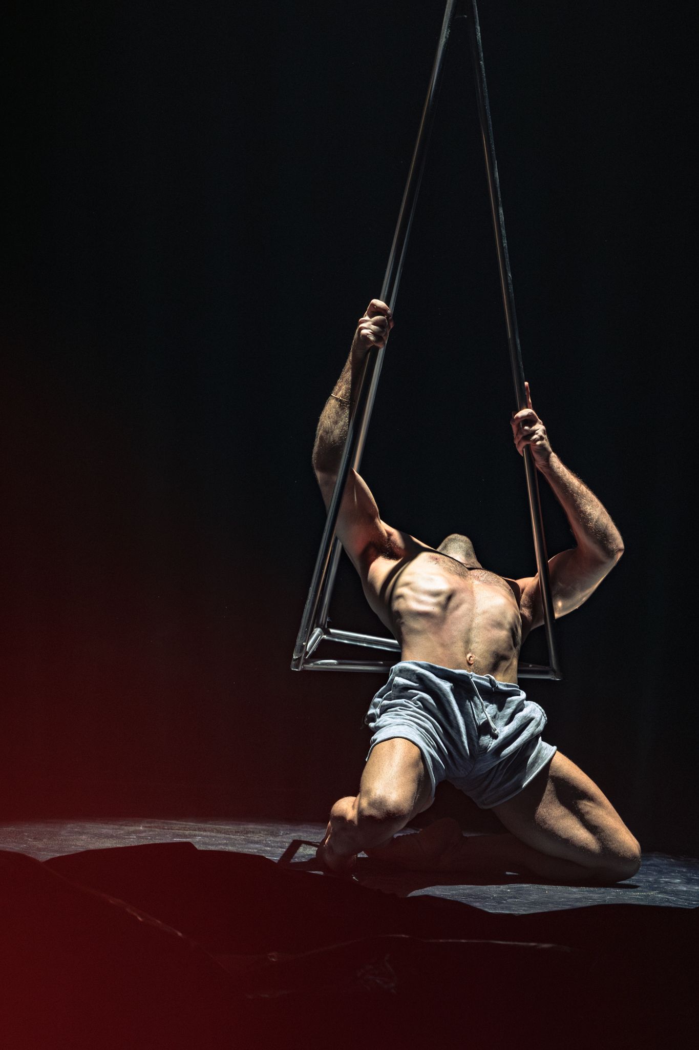 Shirtless aerial performer in denim shorts gracefully suspends from trapeze straps against dark theatrical backdrop.