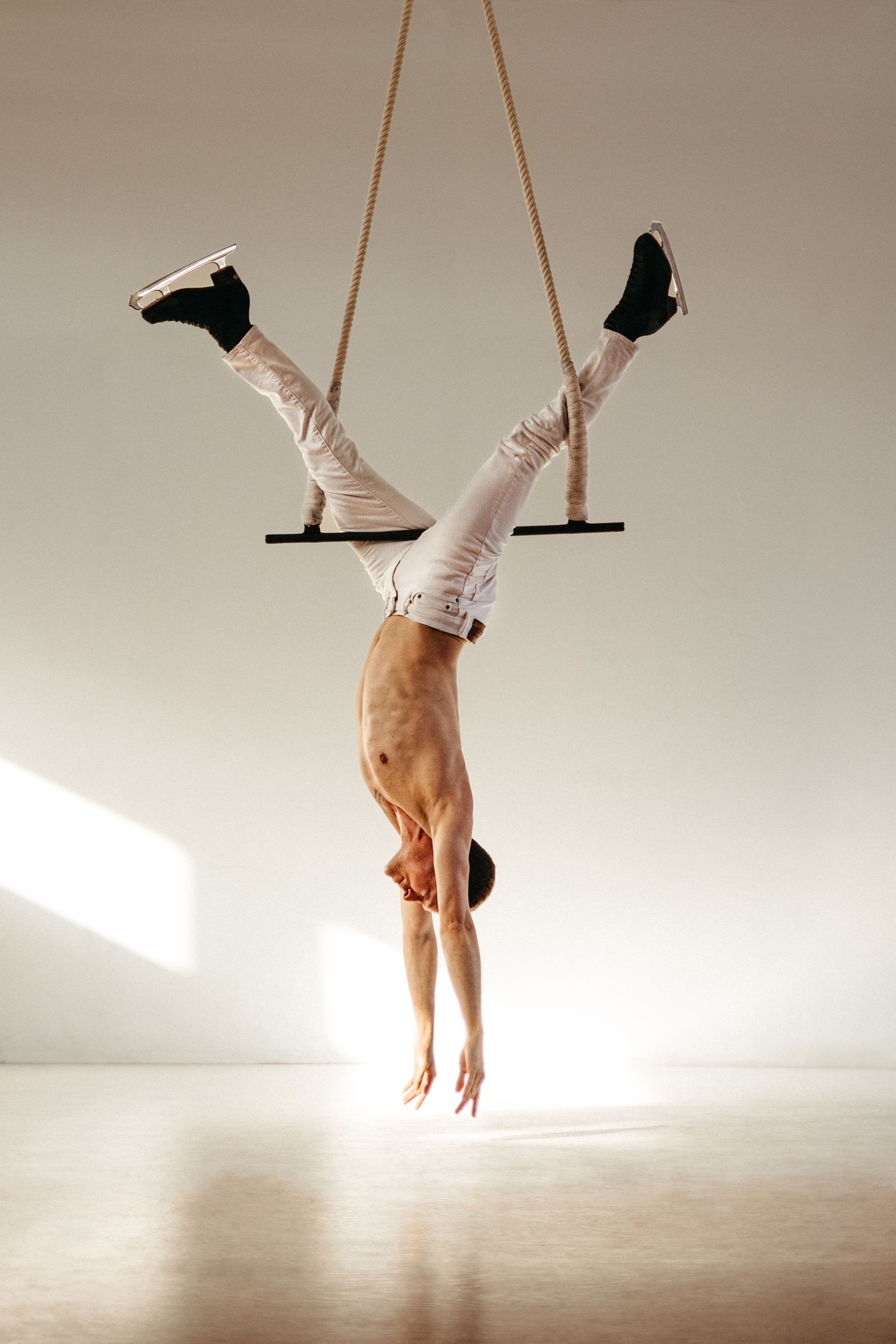 Performer hanging upside down on aerial trapeze wearing white pants and black boots in minimalist setting.