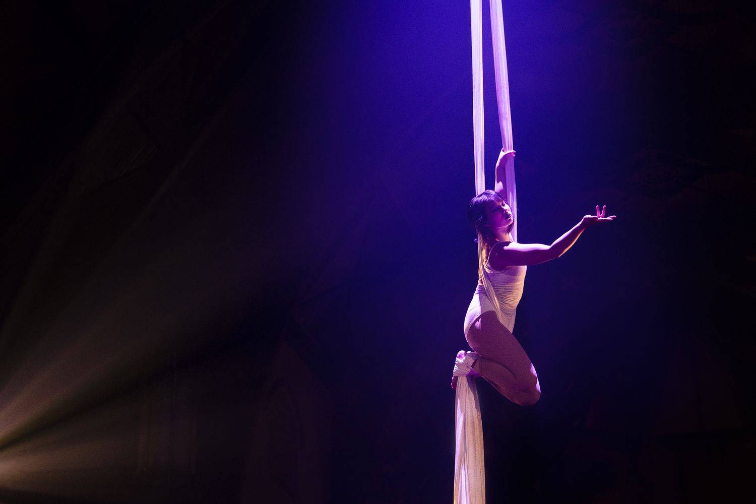 Aerial silk performer executing an elegant split pose against dramatic blue lighting on white fabric.