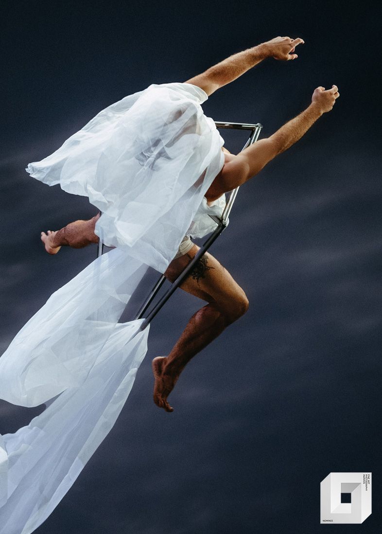 A dancer in flowing white fabric performs a dynamic mid-air leap against a dark background.