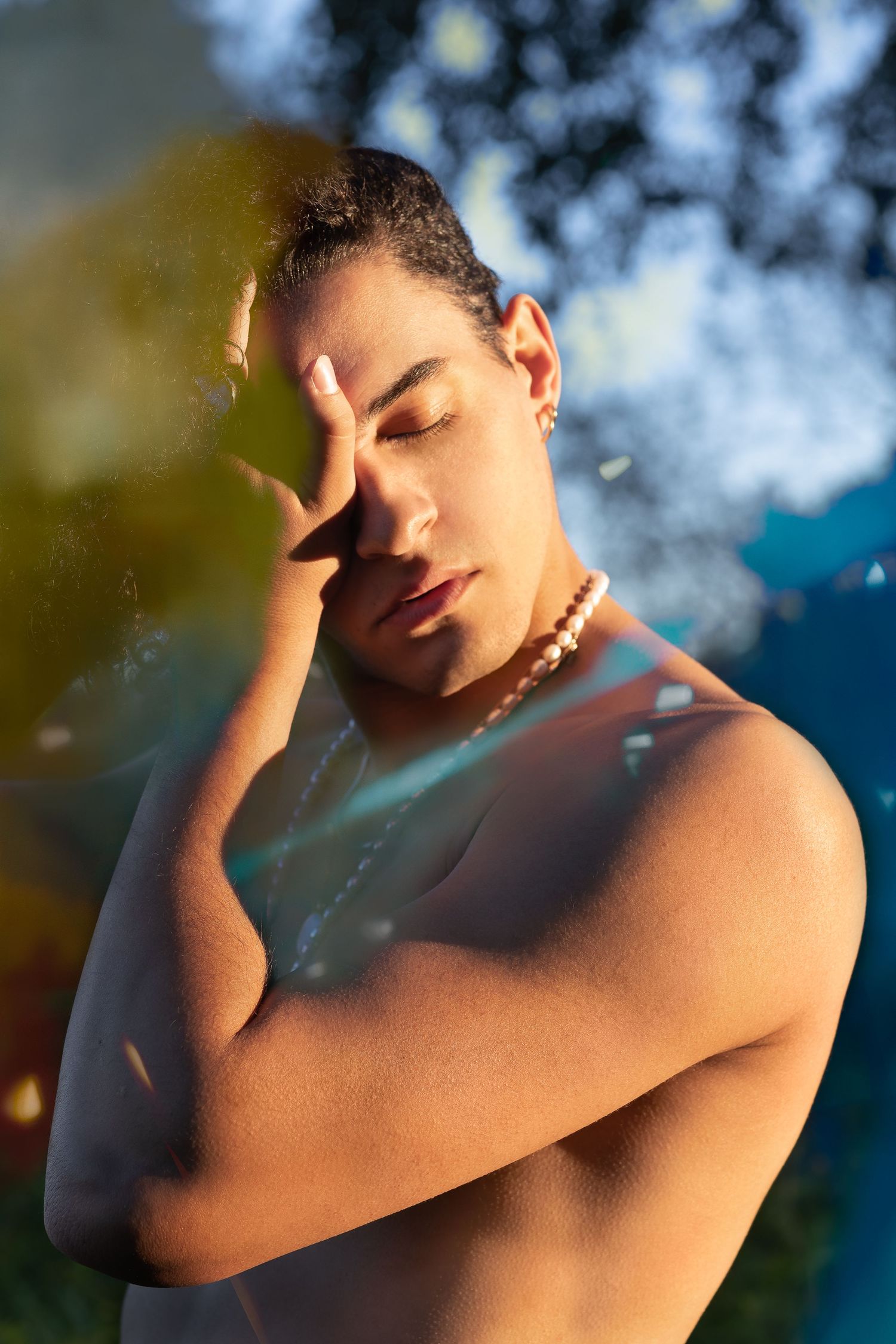 A person poses shirtless outdoors in golden sunlight with their hand raised to their face and eyes closed.