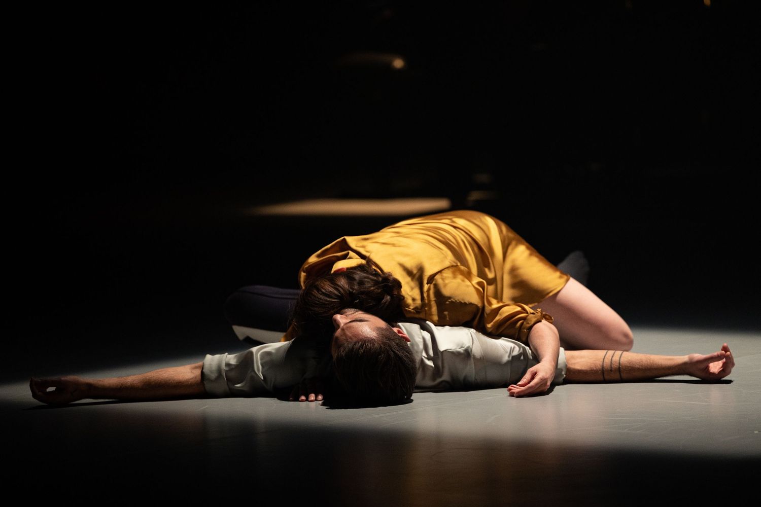 A person in a gold dress lies motionless on a dark stage floor illuminated by dramatic lighting.
