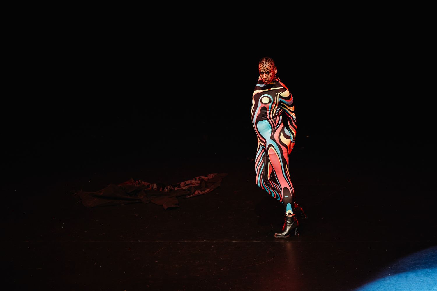 A performer in dramatic body paint and costume stands illuminated on a dark stage in a theatrical performance.