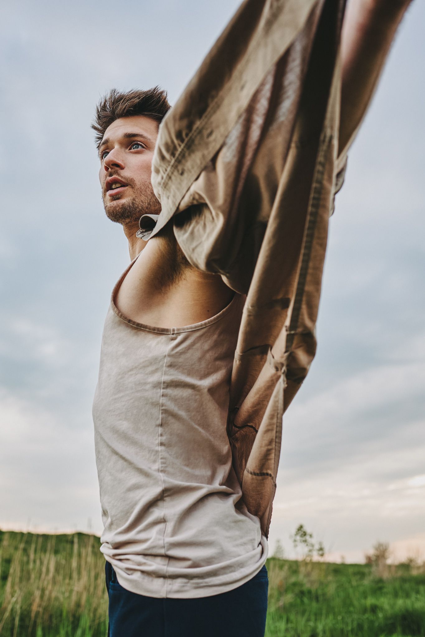 Athletic person stretches outdoors in a beige tank top against a cloudy sky and grassy field.