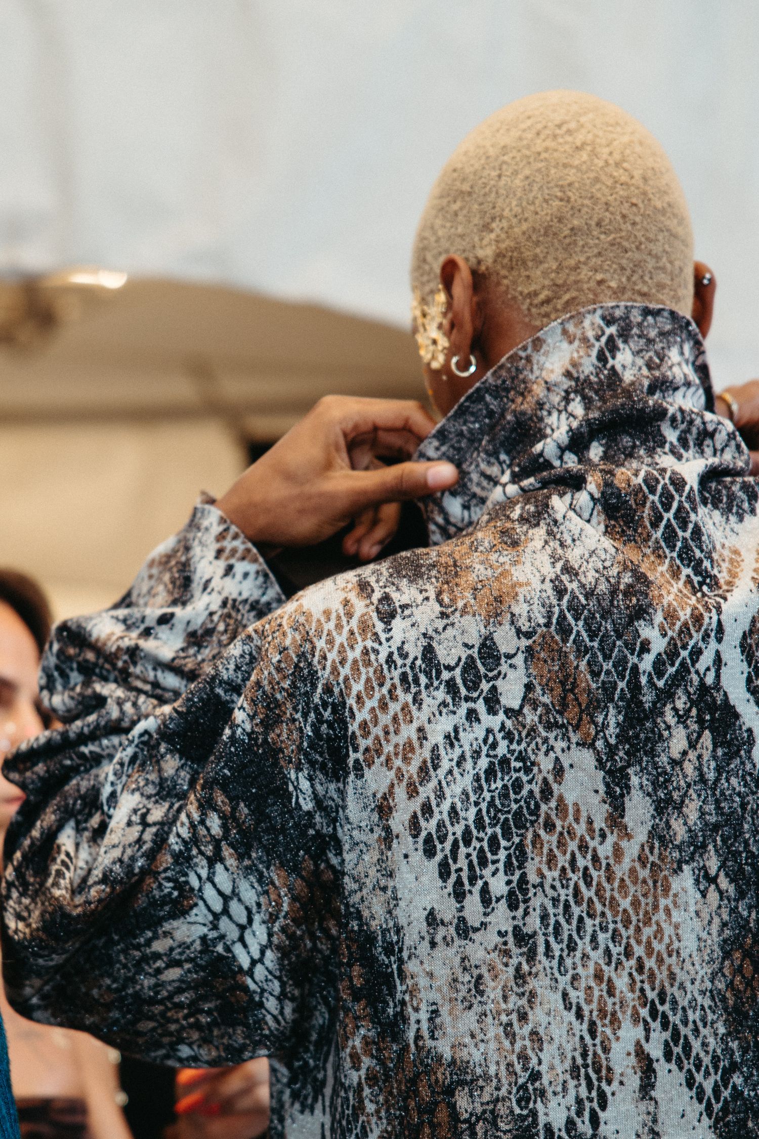 Person wearing a textured blue and white snakeskin pattern jacket with gold ear jewelry and blonde hair.