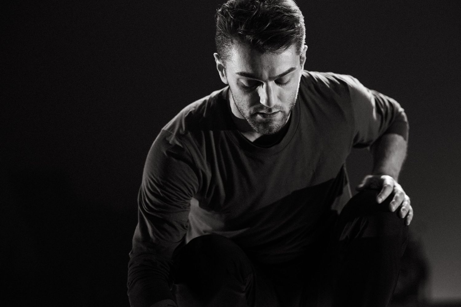 Black and white portrait of a person crouched in a moody, dramatic pose wearing a dark t-shirt.