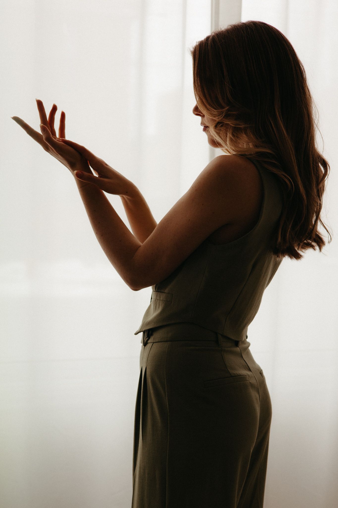 Silhouetted figure stands in elegant pose by window with soft backlight creating dramatic shadows on flowing dress.