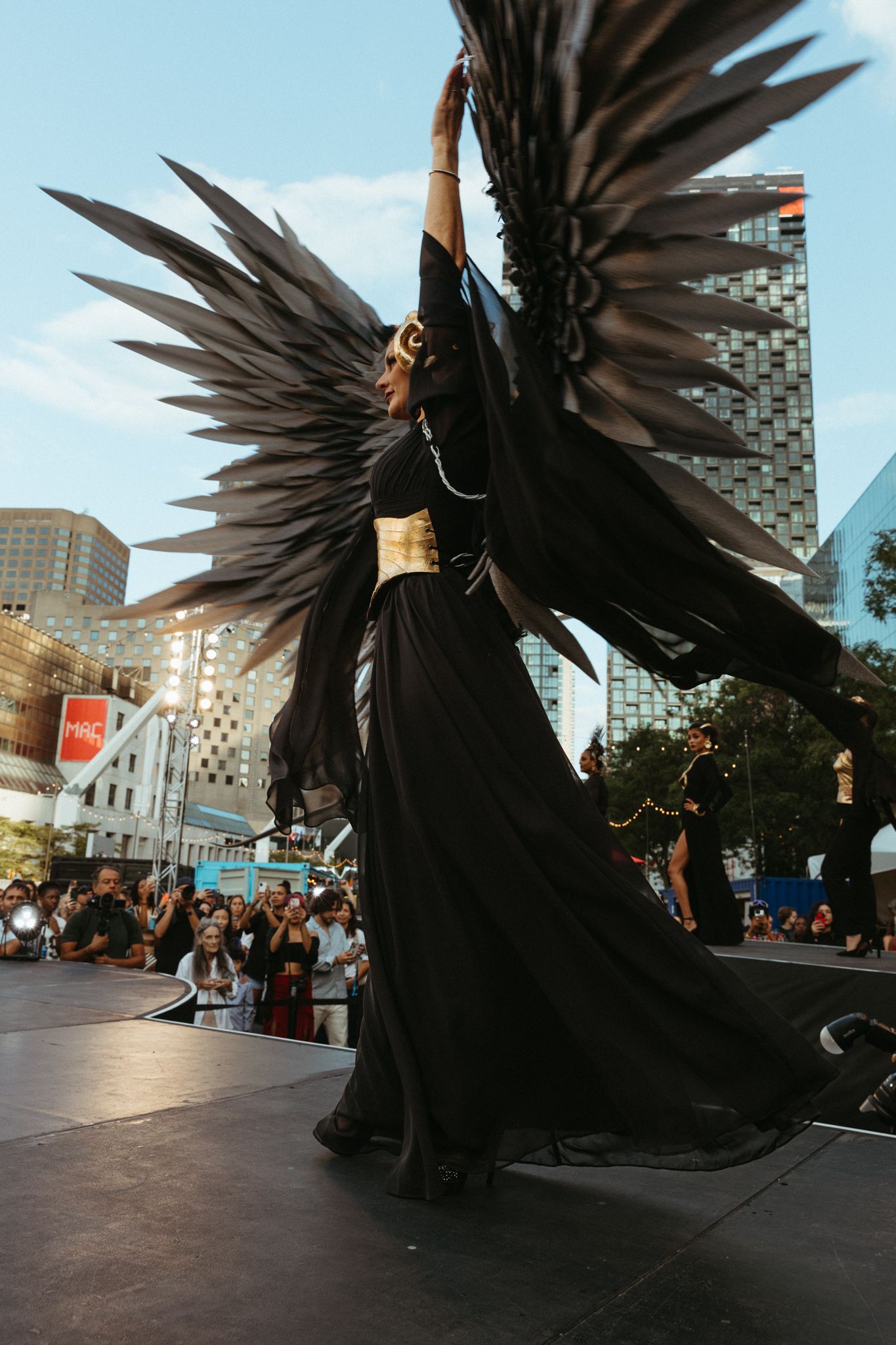 A dramatic figure with large black wings performs on an outdoor stage at an urban plaza during sunset.