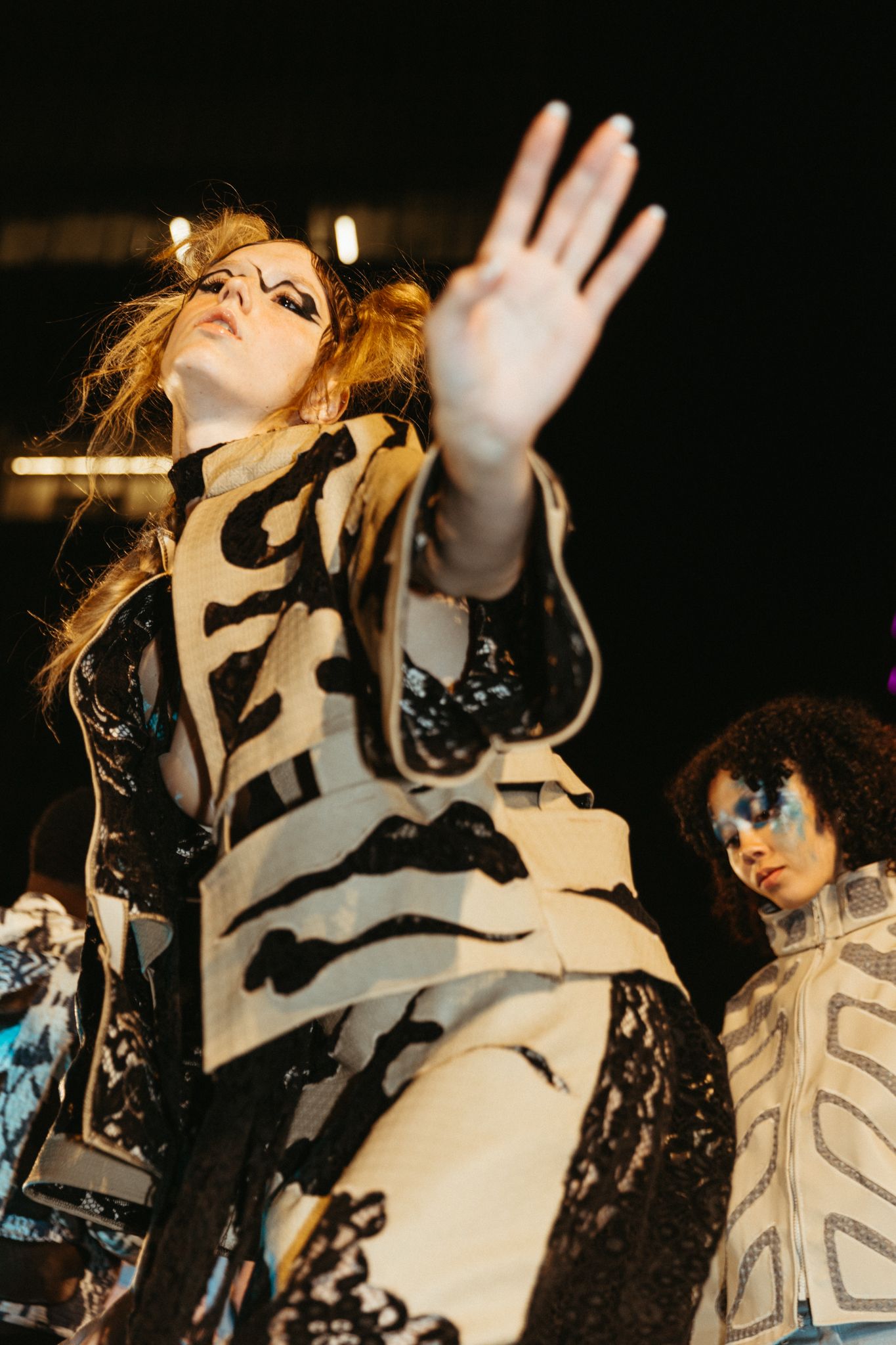 A performer in a dramatic black and white costume raises their hand during an energetic stage performance.