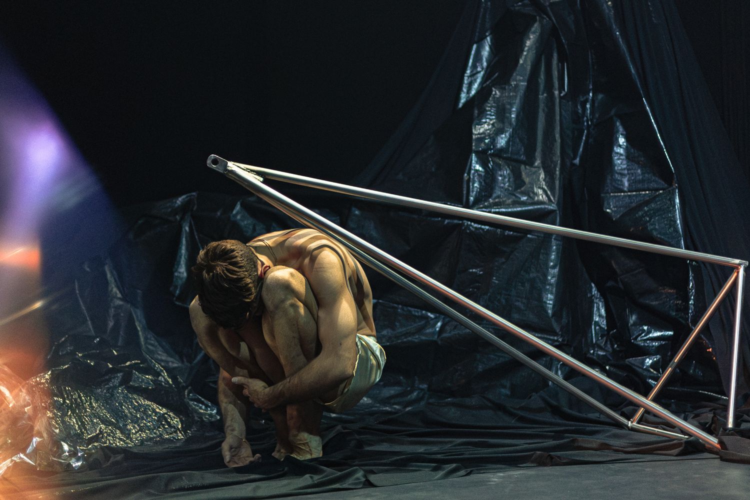 Dramatic performance art piece featuring a crouched performer on stage with metal poles and atmospheric lighting effects.