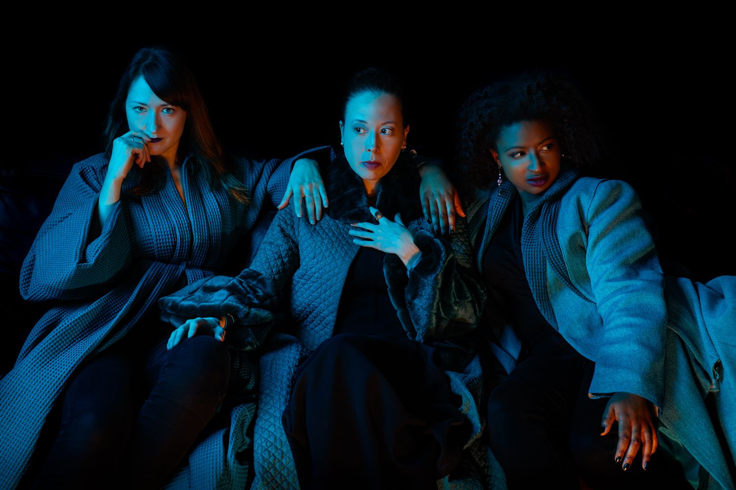 Three figures in dramatic blue lighting sit together on a couch wearing winter coats in an atmospheric scene.