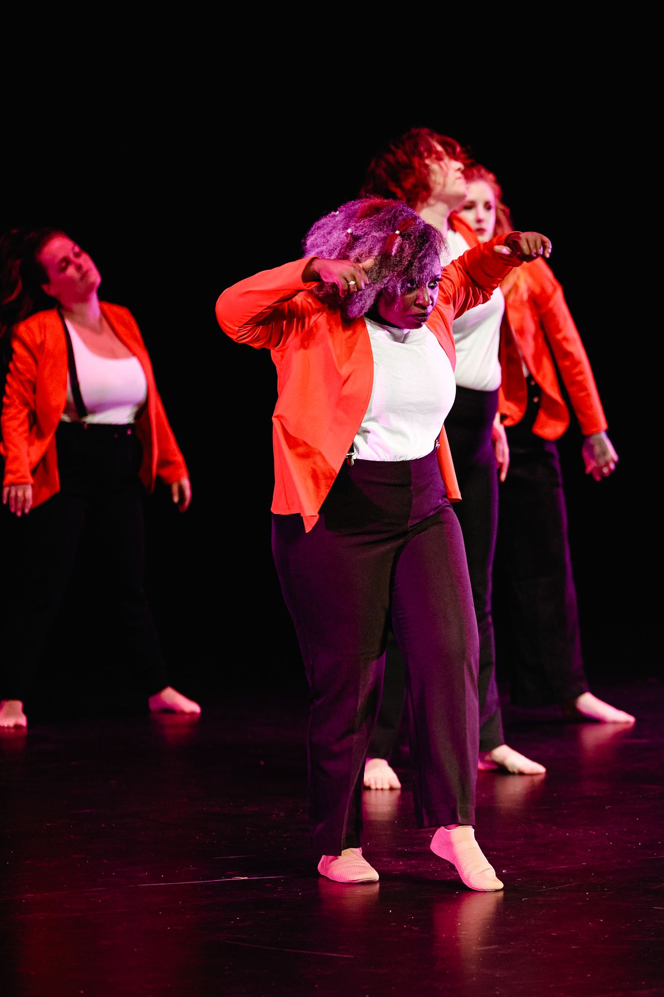 Dancers in red jackets and black pants perform an energetic contemporary dance routine on a dark-lit stage.