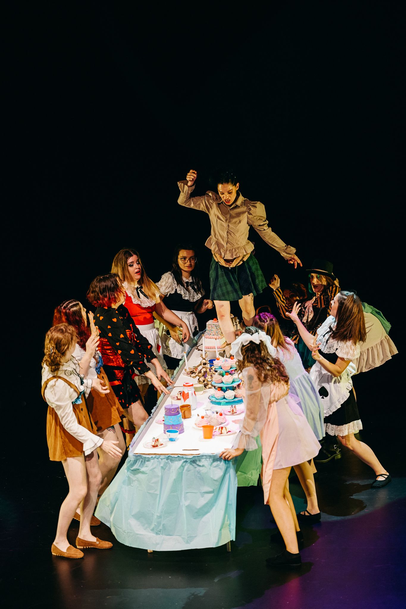 A theatrical scene shows performers gathered around a dining table in dramatic poses during what appears to be a chaotic party moment.