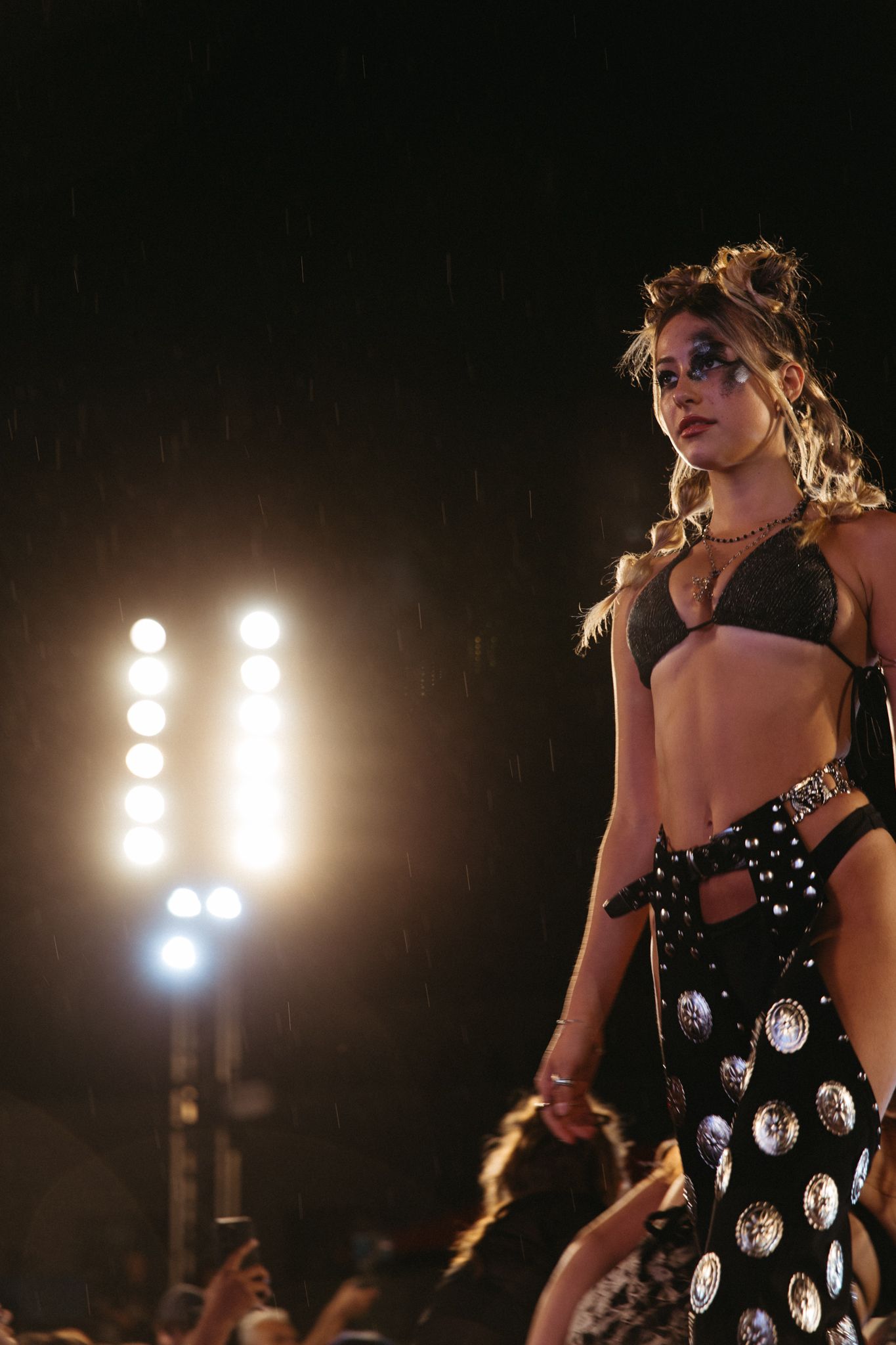 Model walks runway in black bikini with silver medallion skirt under dramatic stage lighting at fashion show.