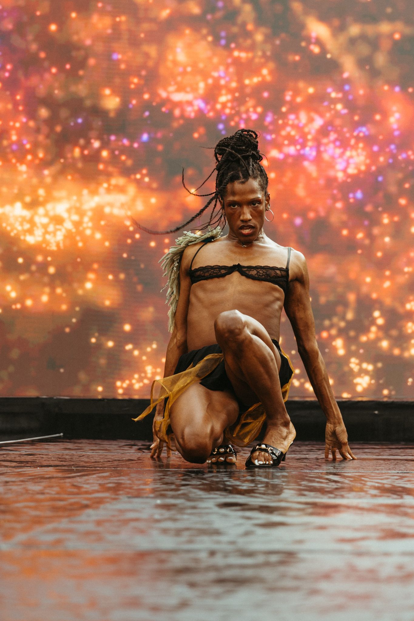 A dancer performs in shallow reflecting water against a backdrop of vibrant orange and pink sparkles and lights.