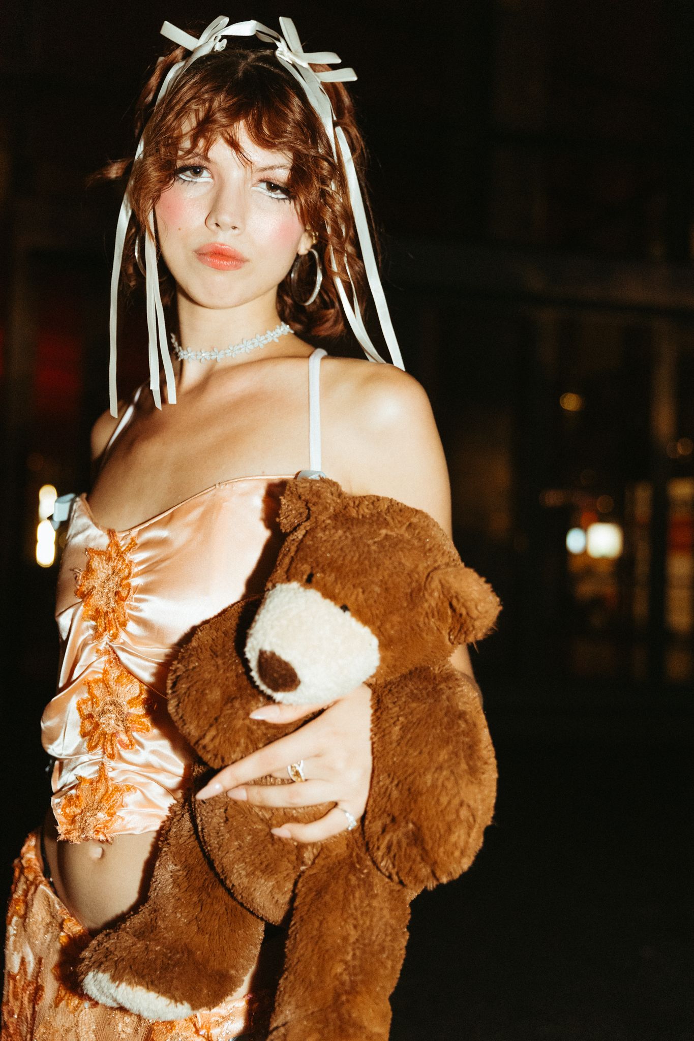 A woman poses at night in a teddy bear-like brown fur coat with white ribbons in her hair.
