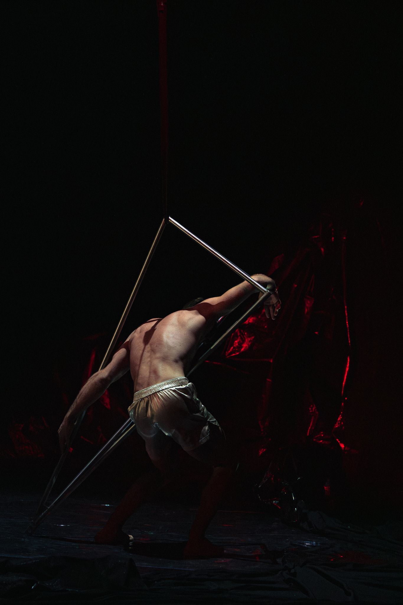 Dramatic dark artistic photo of shirtless performer doing pole acrobatics with red lighting effects in background.