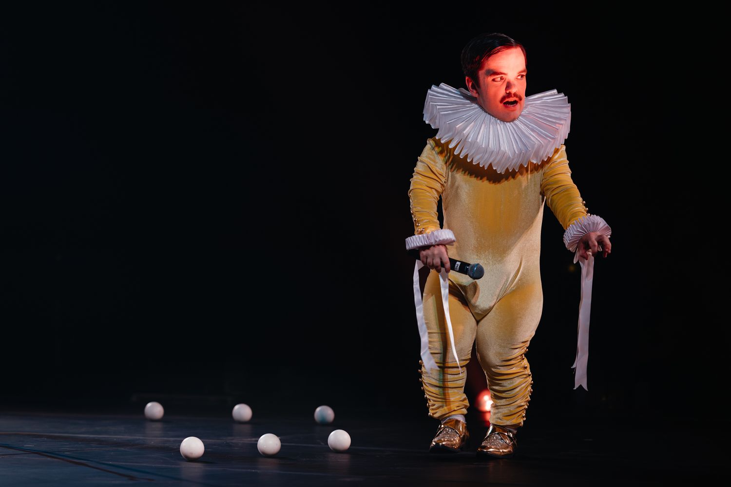 Performer in yellow Elizabethan-style costume with white ruffled collar hunches over juggling balls on dark stage.