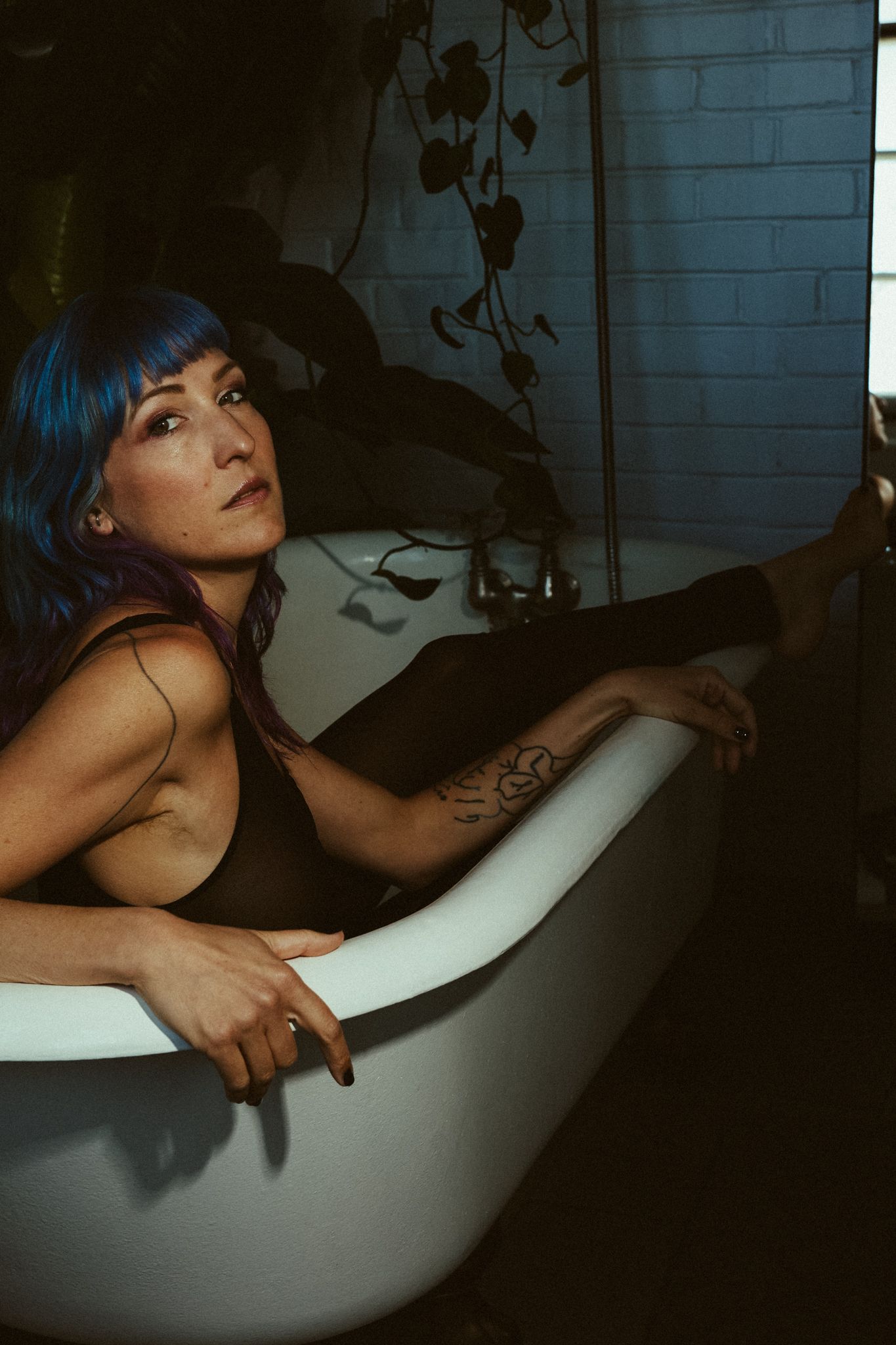 A moody boudoir photo in a bathtub with blue hair and dark shadows against white porcelain.