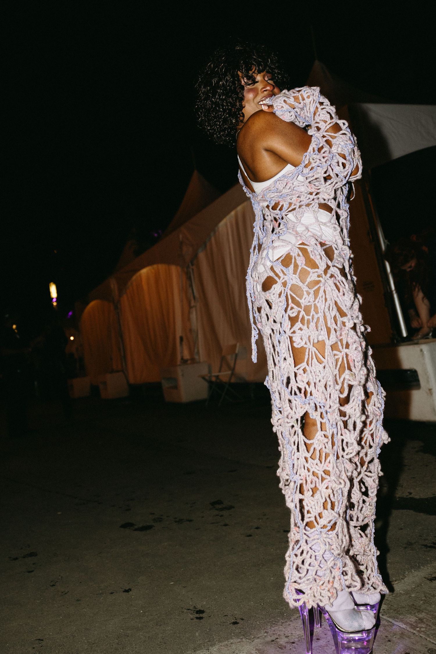 A figure in a white crochet lace dress and purple platform boots poses against a dark background at night.