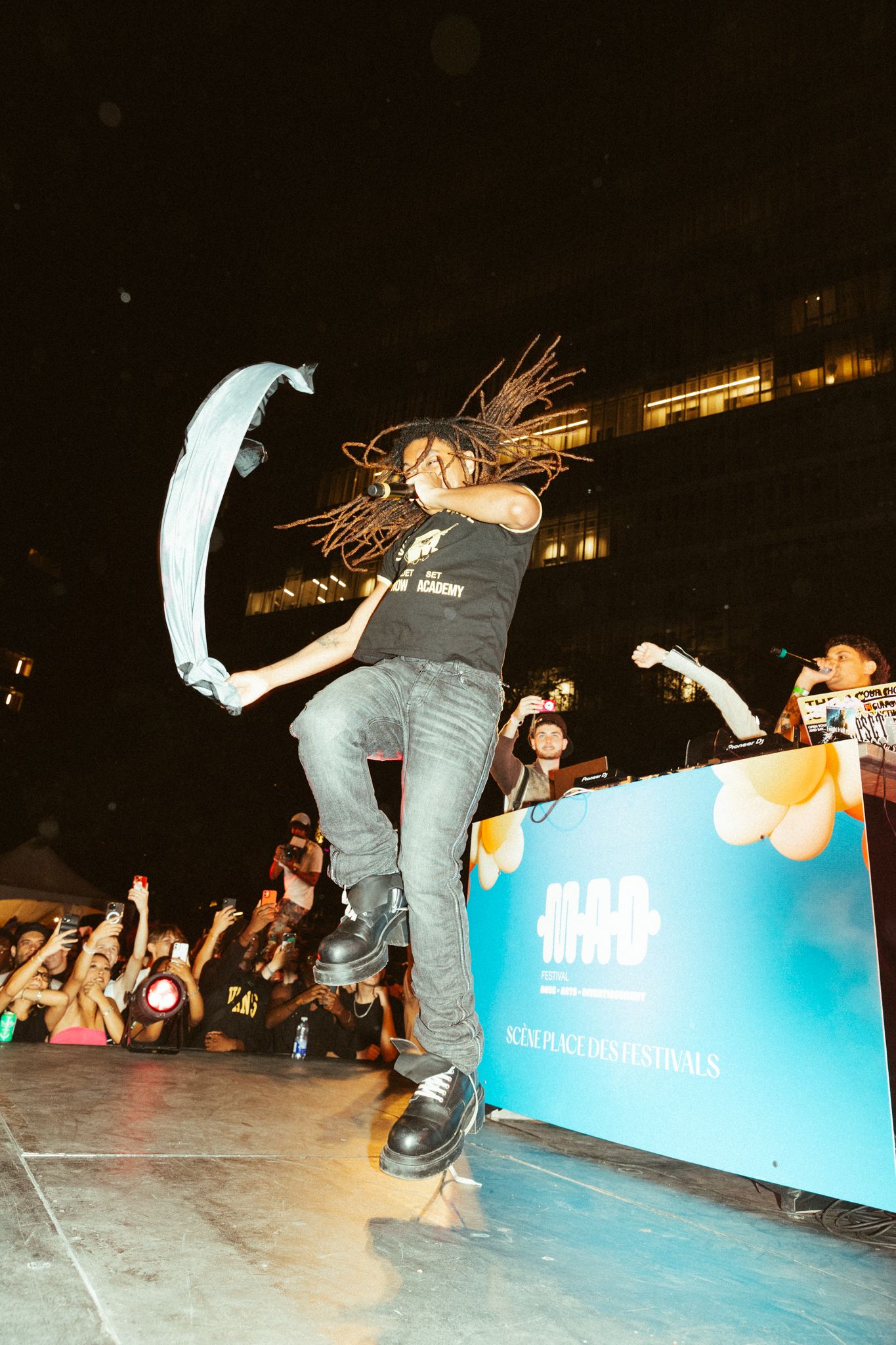 An energetic performer on stage with dreadlocks jumping during a concert with crowd and illuminated backdrop.