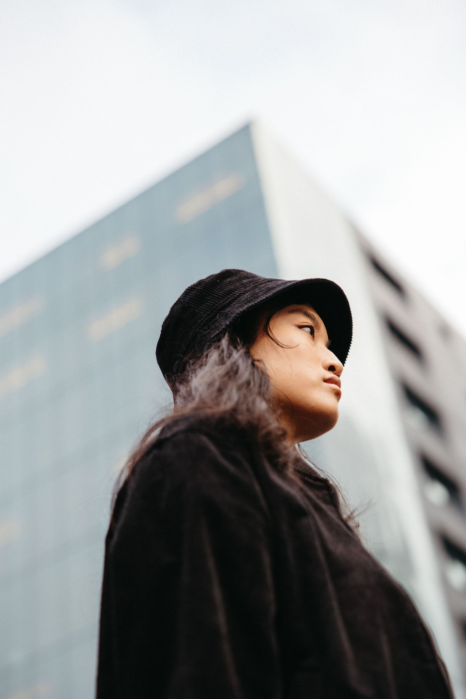 A figure in a dark coat and cap gazes upward against an urban backdrop of buildings.