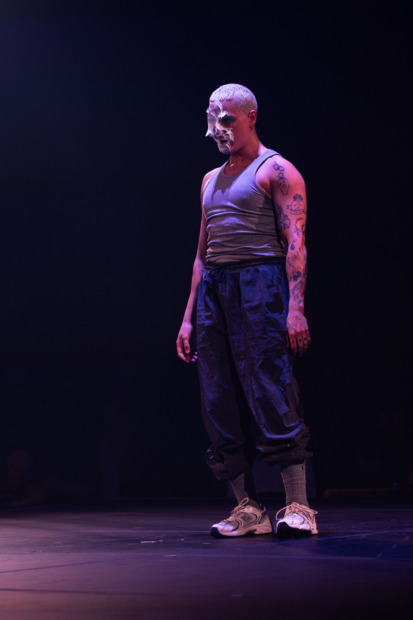 A performer in dark clothing stands on stage under moody theatrical lighting during a dramatic dance performance.