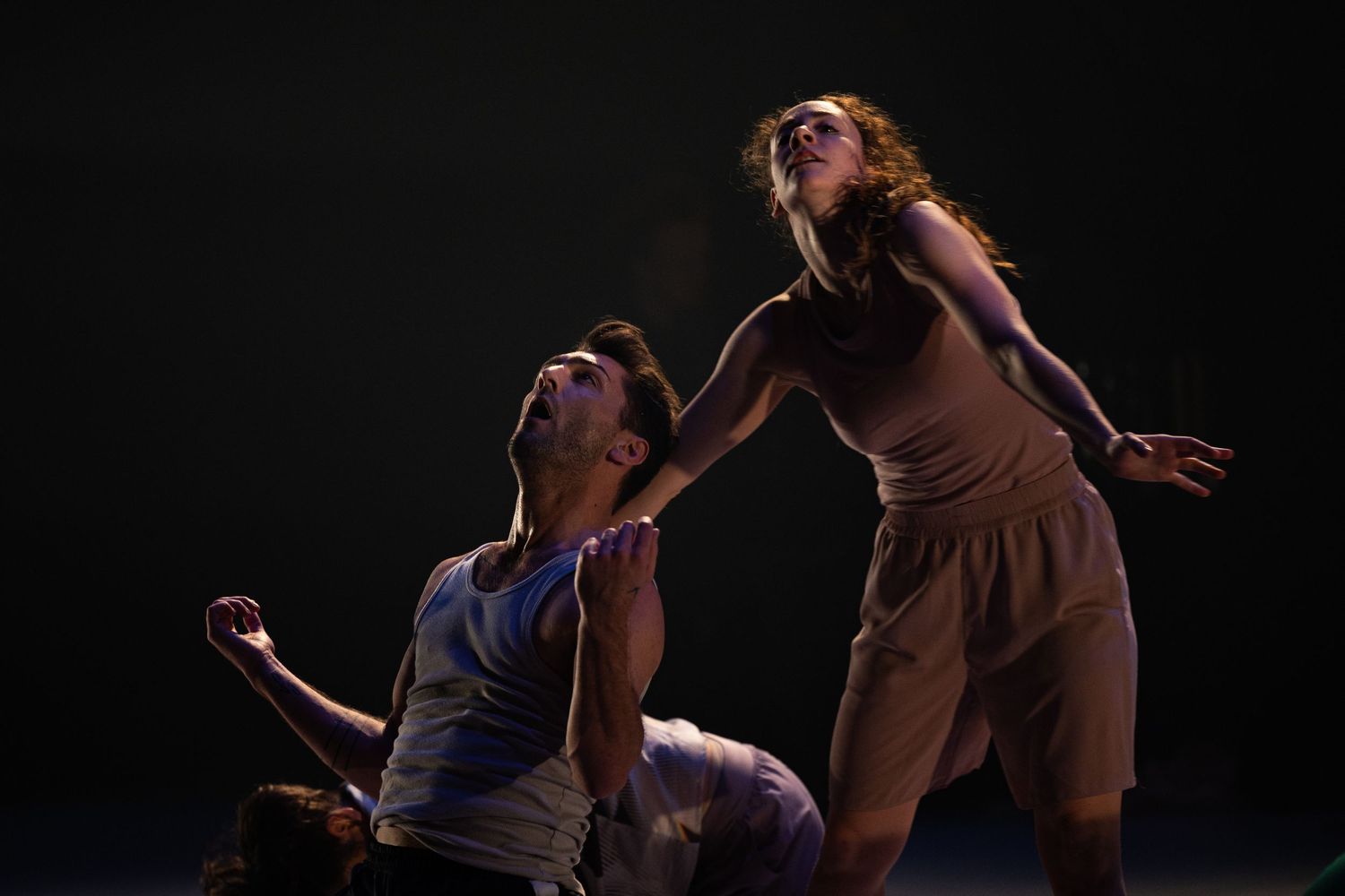 Two modern dancers perform an emotional duet on a dark stage, their bodies creating dramatic shapes in the lighting.
