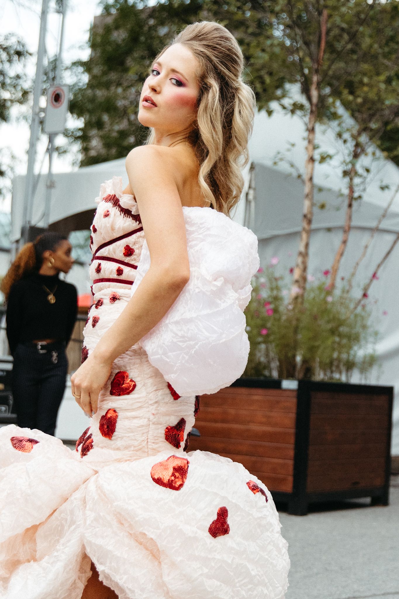 A person in a white ruffled dress with red heart accents twirls in an outdoor setting near trees and benches.