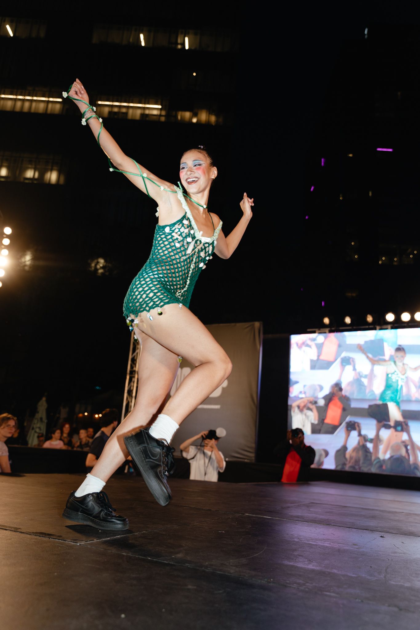 Performer in a sparkling teal leotard and white boots strikes an energetic dance pose on a dark stage.