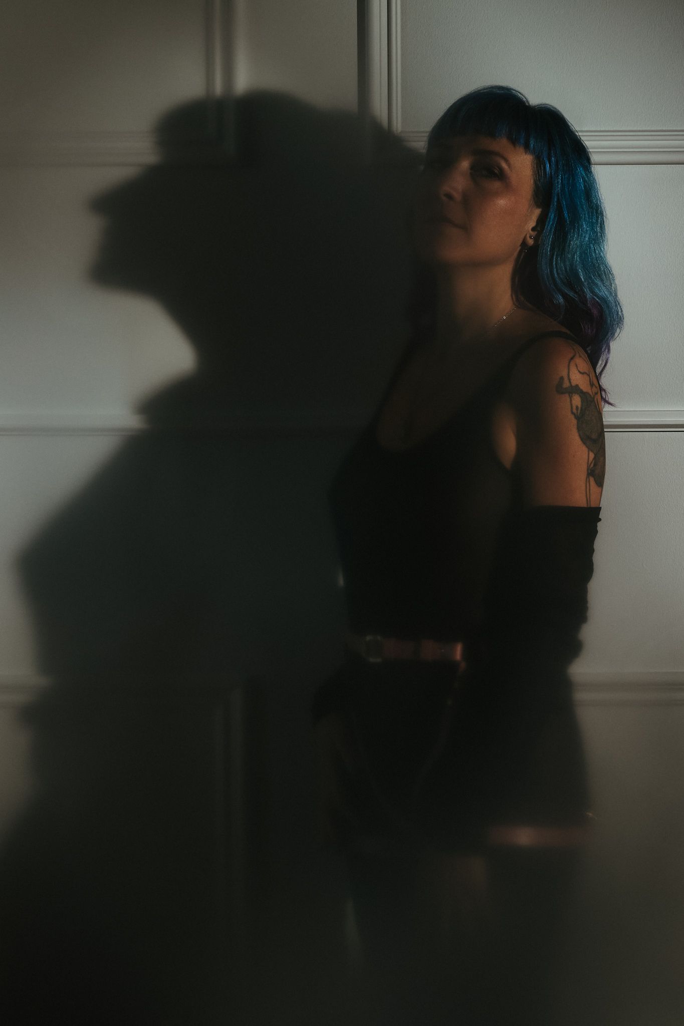 Dark moody portrait with blue-haired figure casting dramatic shadow against white wall in dim lighting.