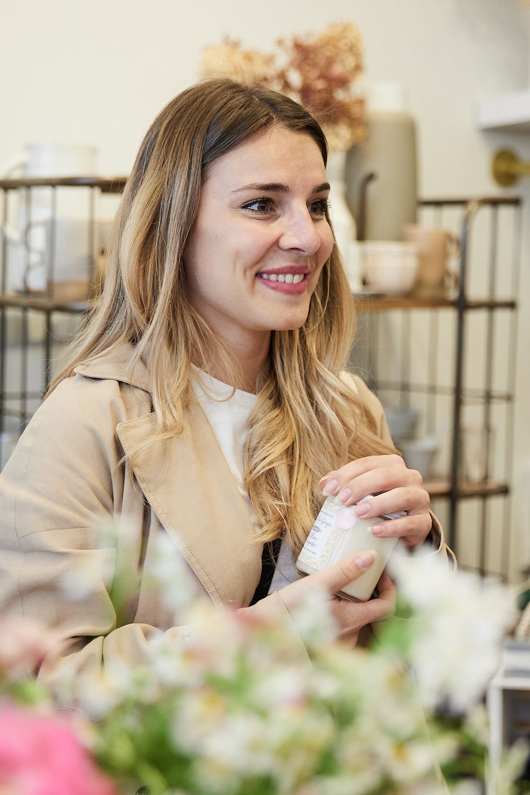 Smiling individual in a beige coat enjoys a beverage while sitting at a table with floral decorations.