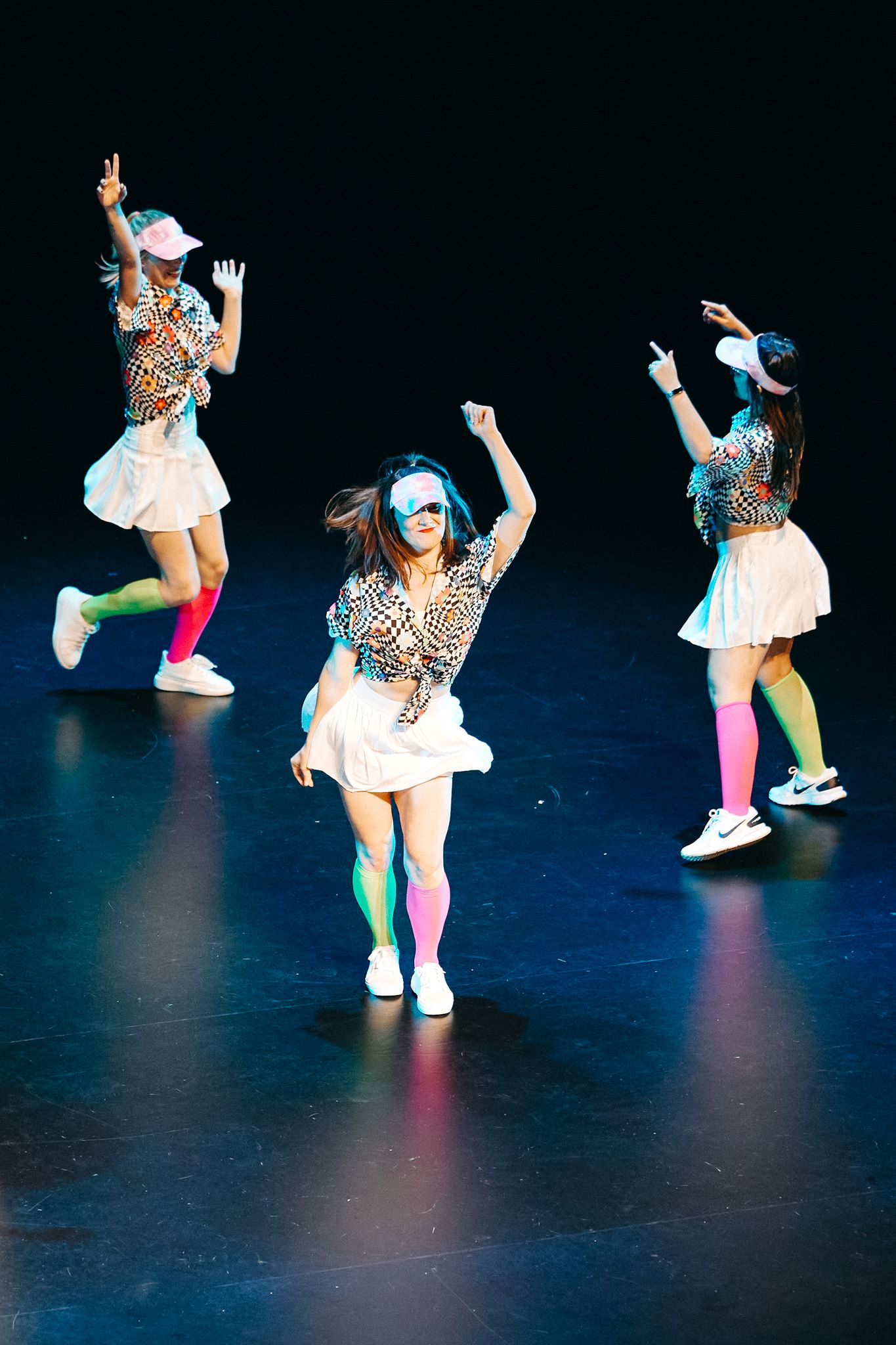 Dancers perform in colorful patterned tops, white skirts and neon socks during an energetic stage performance.