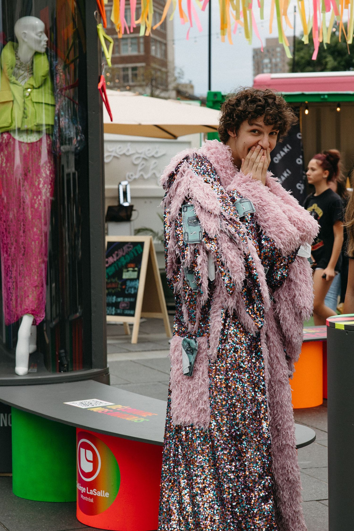 Street scene with a display mannequin in green and pink garments next to someone wearing a sparkly floral dress and pink scarf.