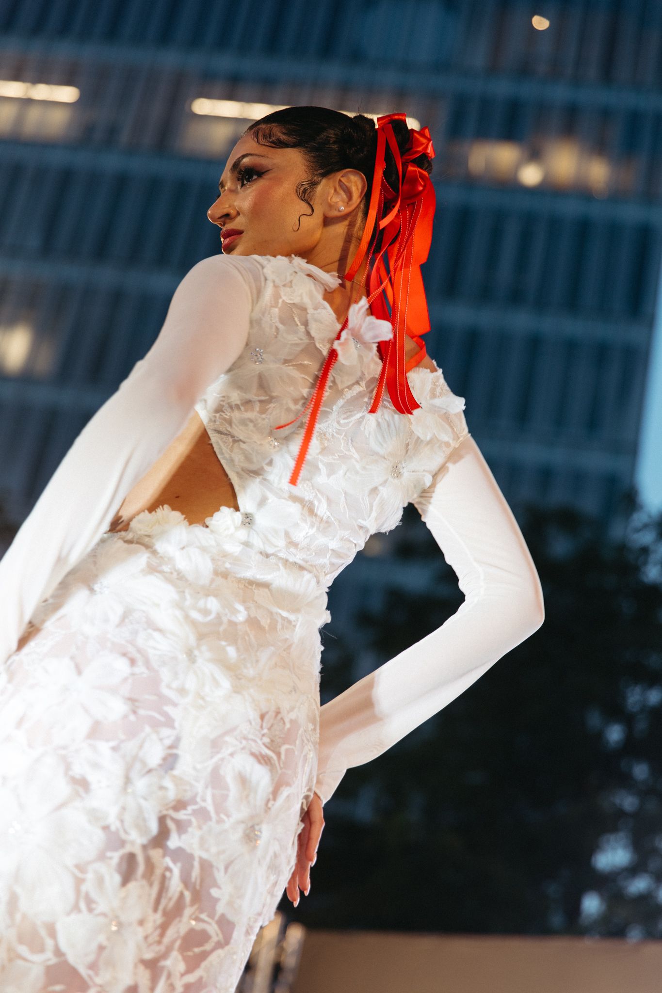 Model in white lace bridal gown with long sleeves and red ribbon hair accessory poses at fashion show.
