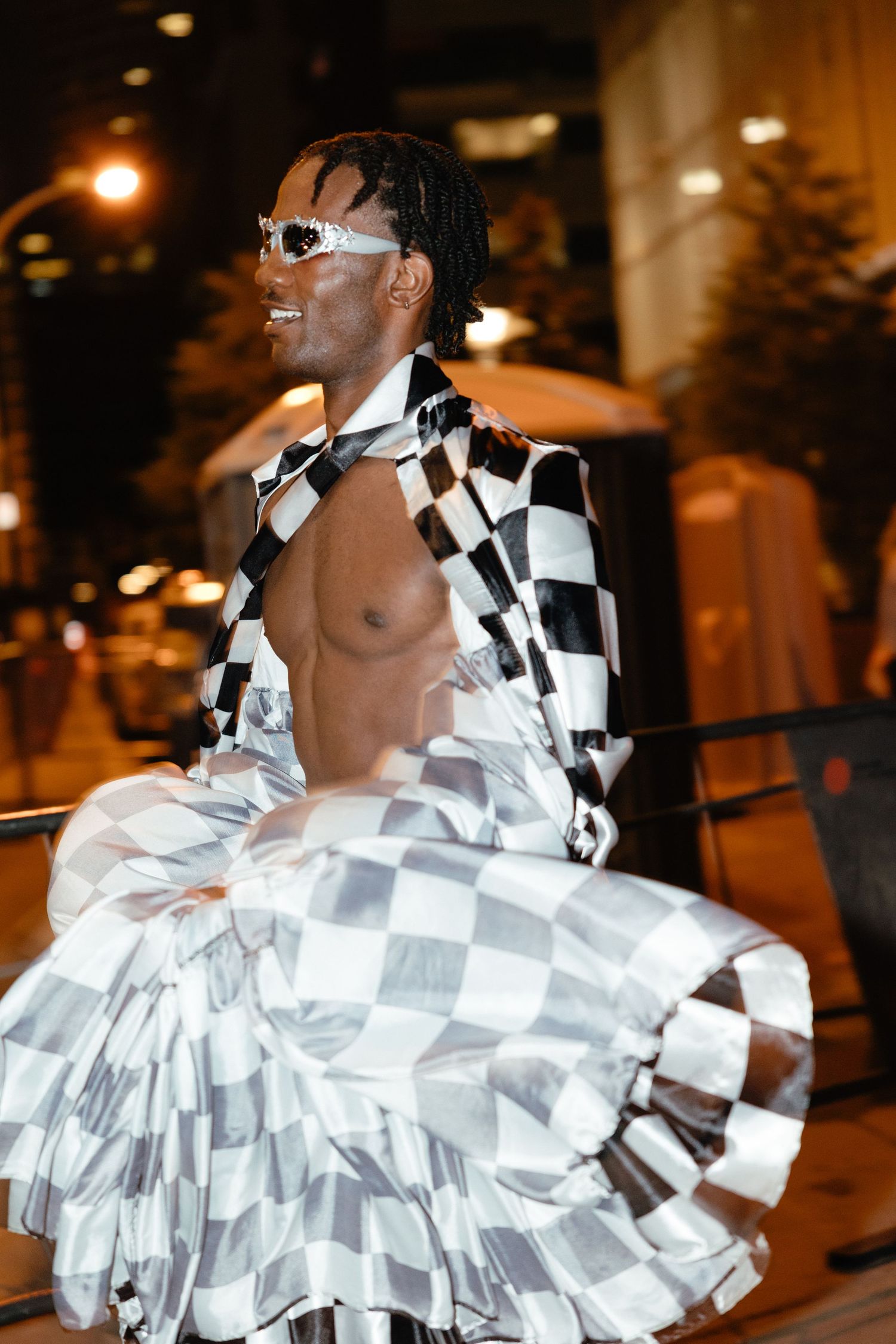 Performer wearing white sunglasses and black-and-white checkered outfit dances outdoors at night under warm lighting.