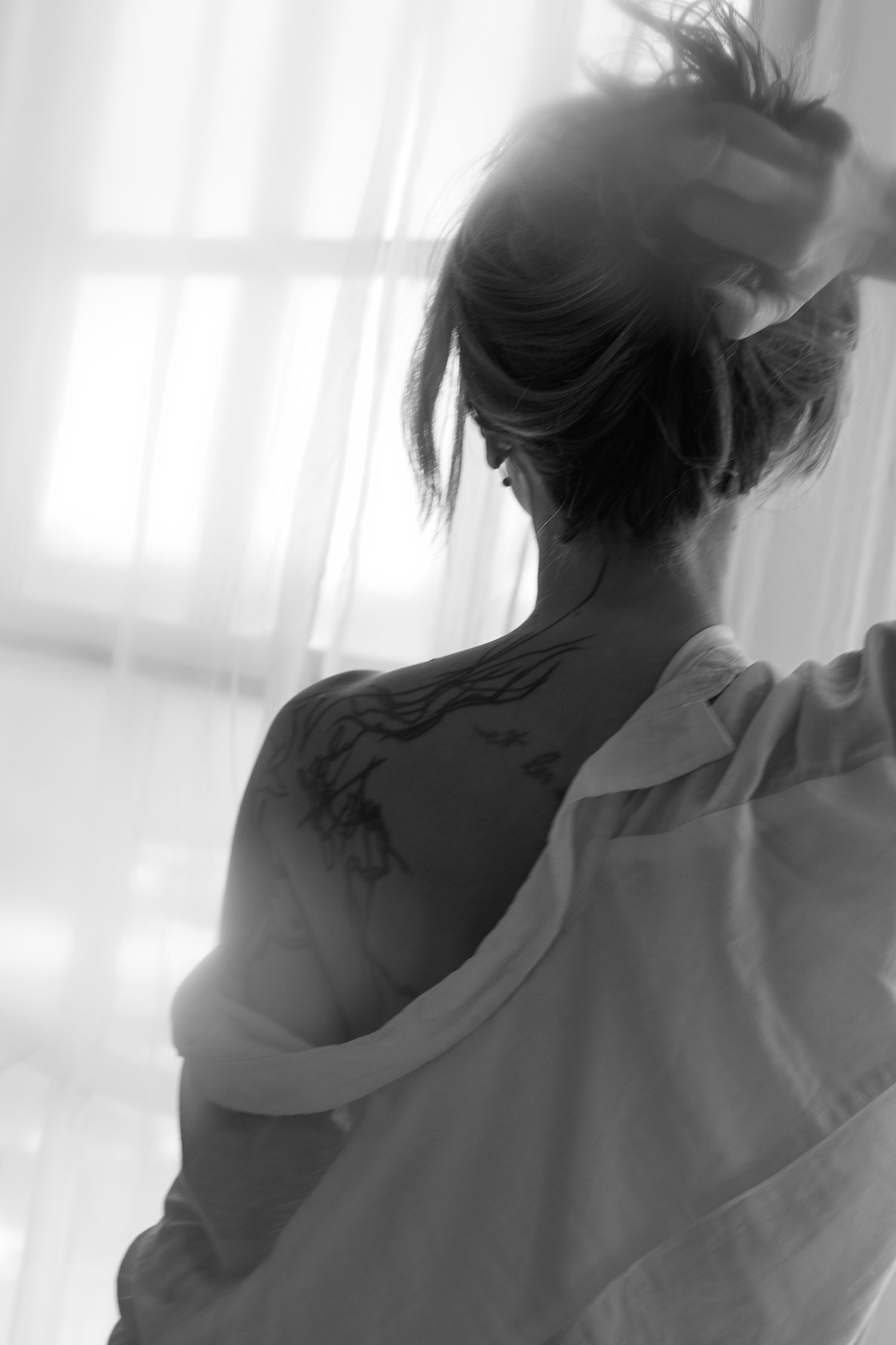 Black and white artistic photo showing silhouette of person from behind with pulled up hair and exposed shoulder in soft light.