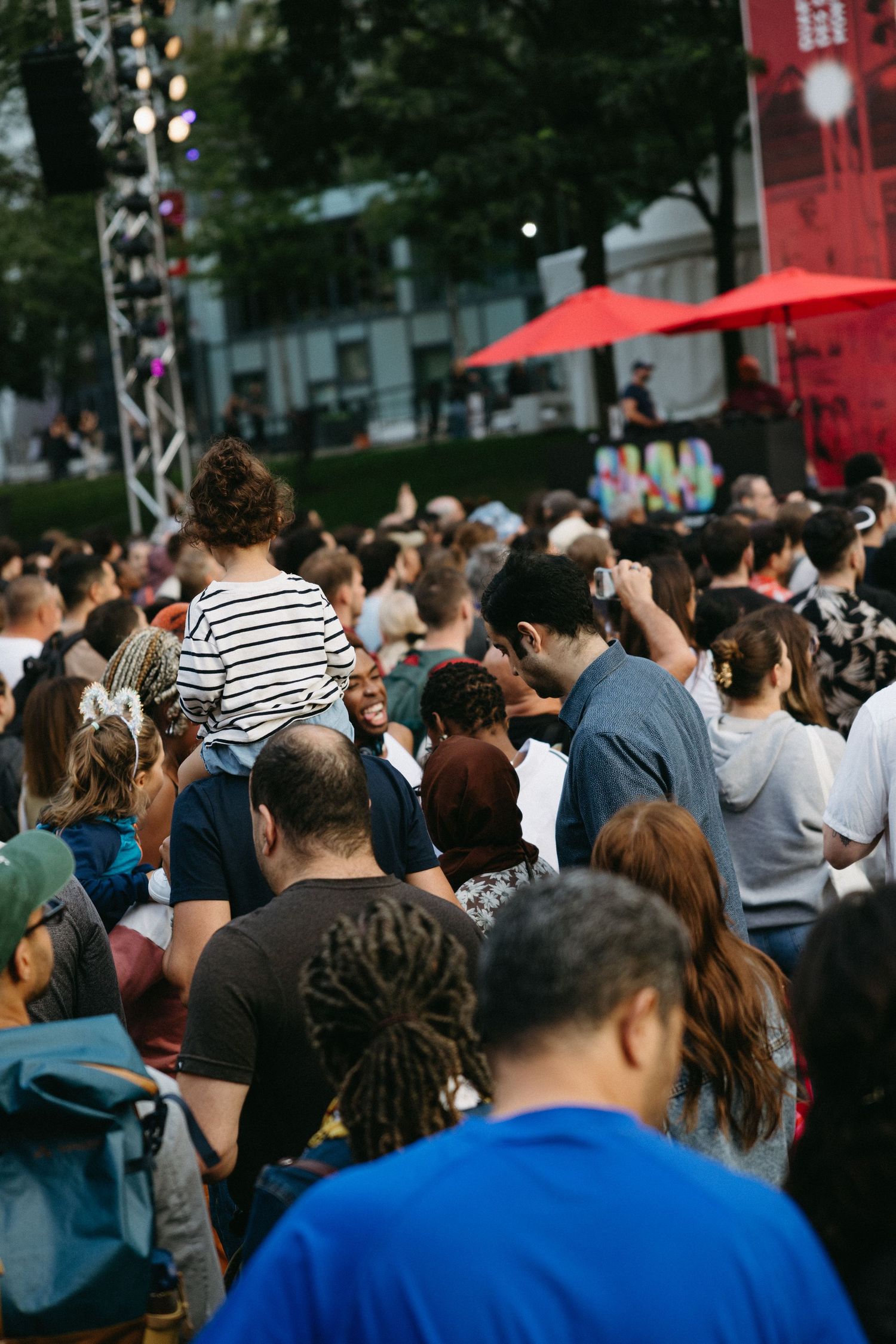 Large crowd of festival attendees gathered outdoors with red umbrellas and stage equipment visible in background.