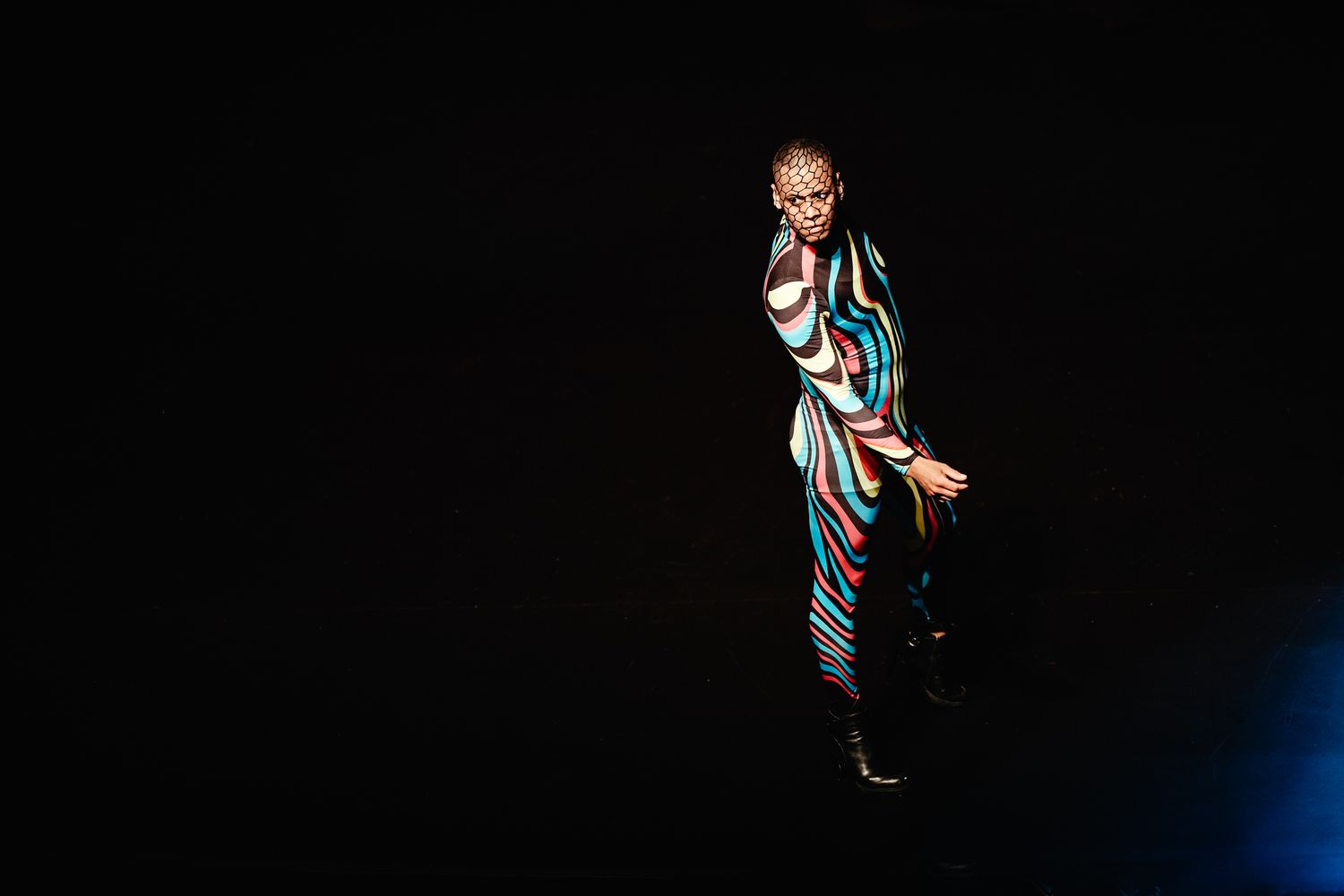 A performer in a striped outfit stands dramatically in a spotlight against a dark background.