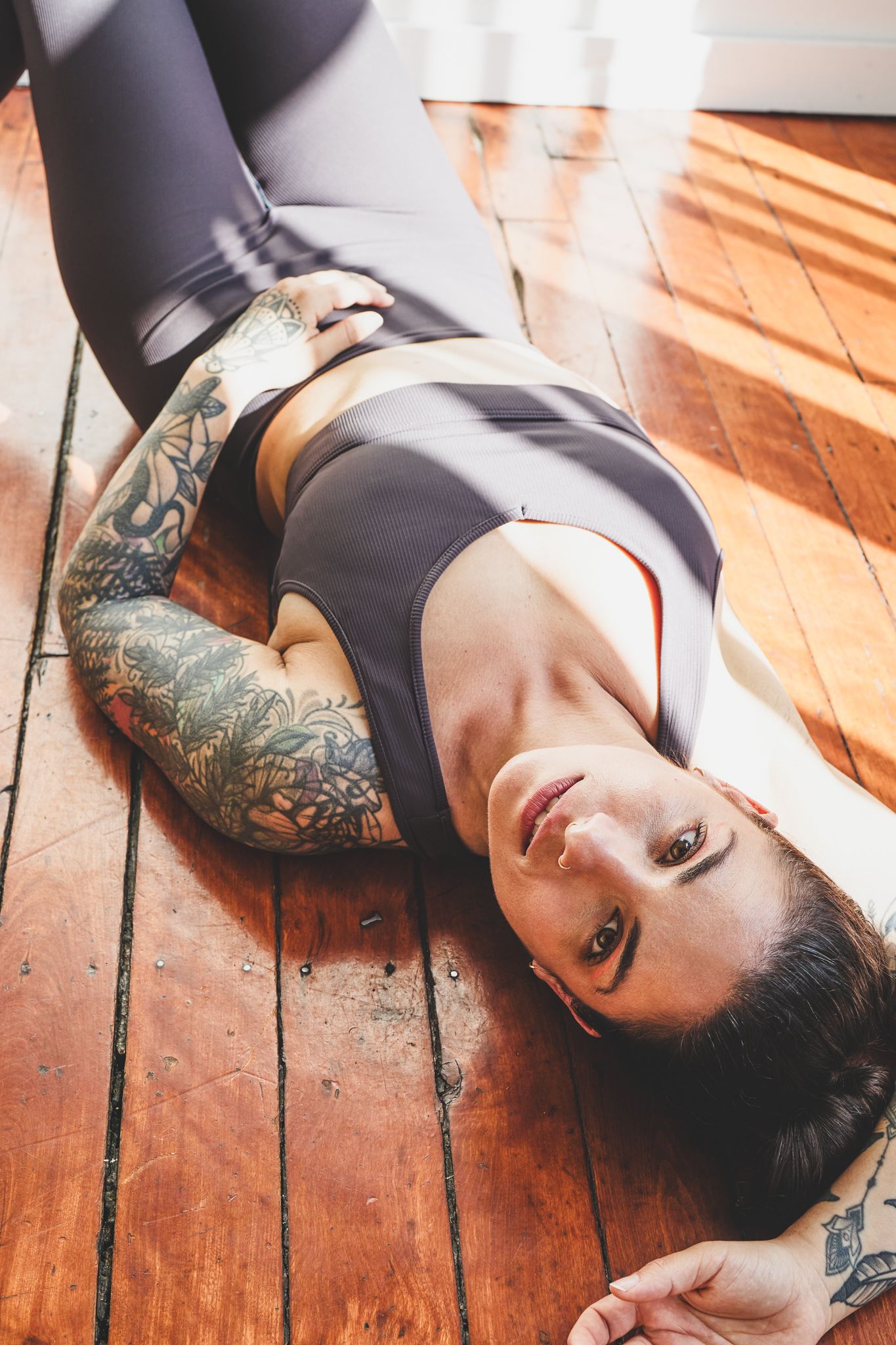 Person lying on wooden floor wearing black athletic wear with visible arm tattoos in natural sunlight.