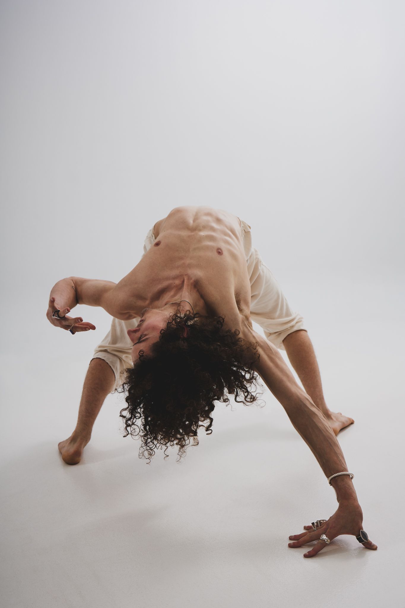 Dancer in beige clothing performs expressive contemporary dance move with curly hair falling back against white background.
