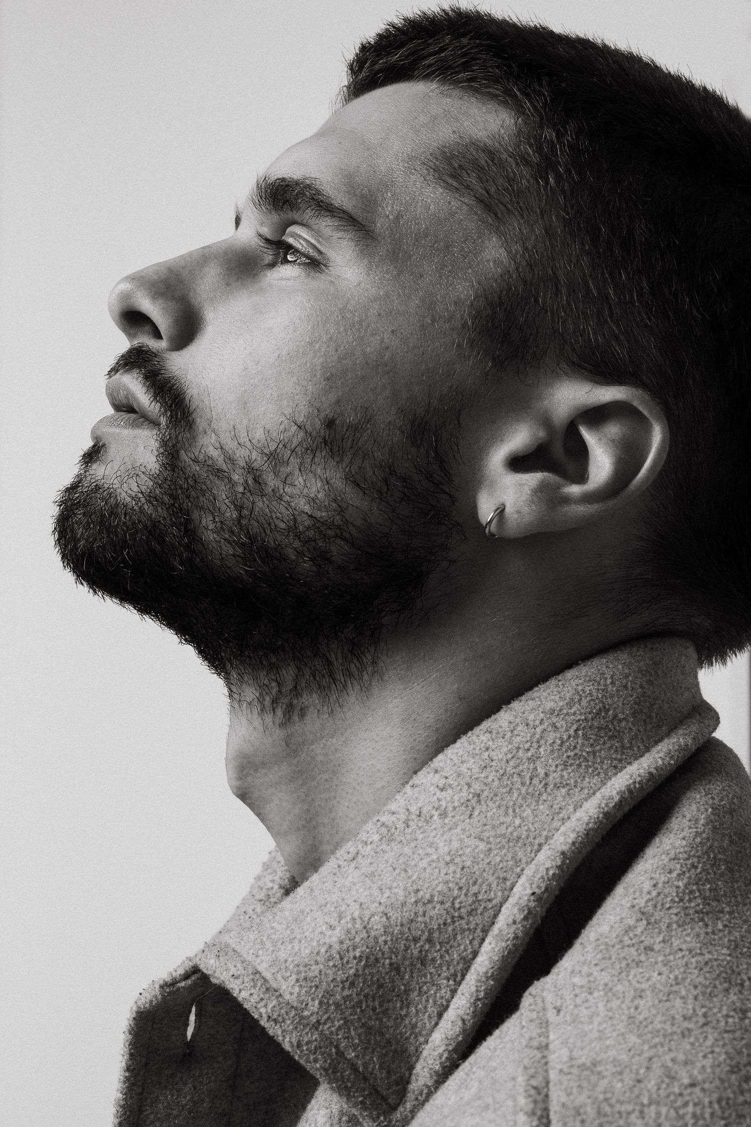 Black and white profile portrait of a bearded man in an upturned collar coat looking upward with dramatic lighting.