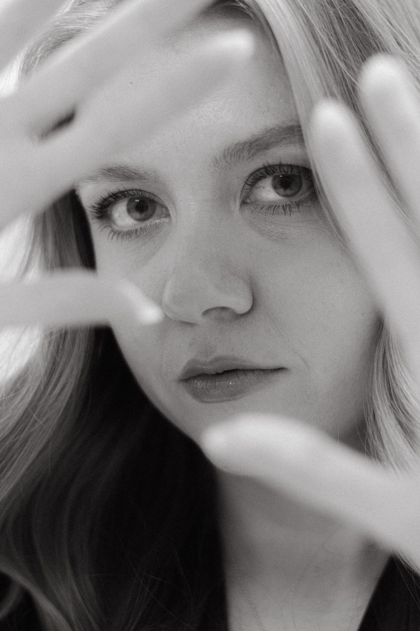 Close-up black and white portrait of a woman peeking through fingers in an artistic pose with soft lighting.