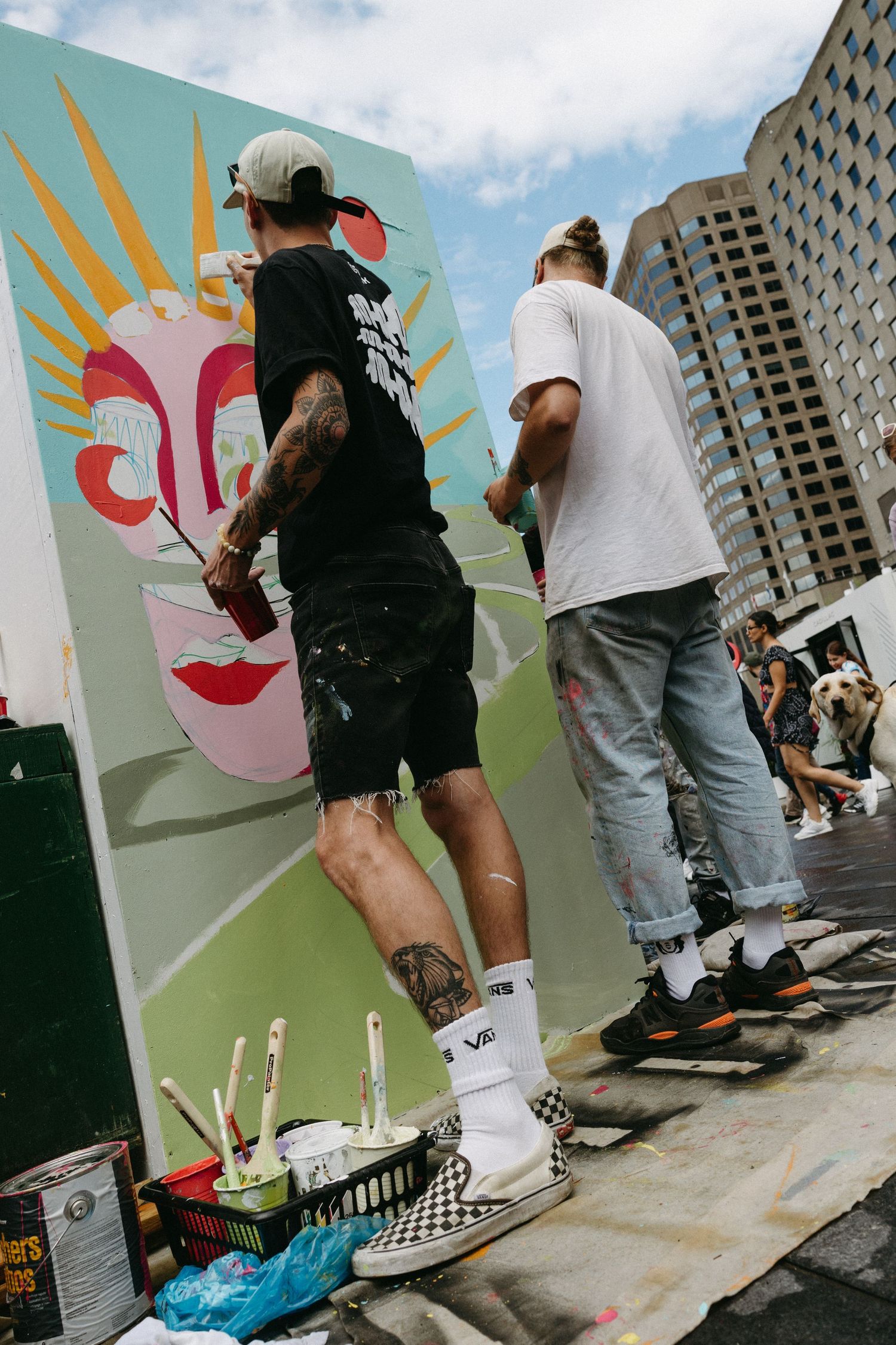 Two street artists work together on a vibrant mural featuring bright sunburst designs in an urban setting.
