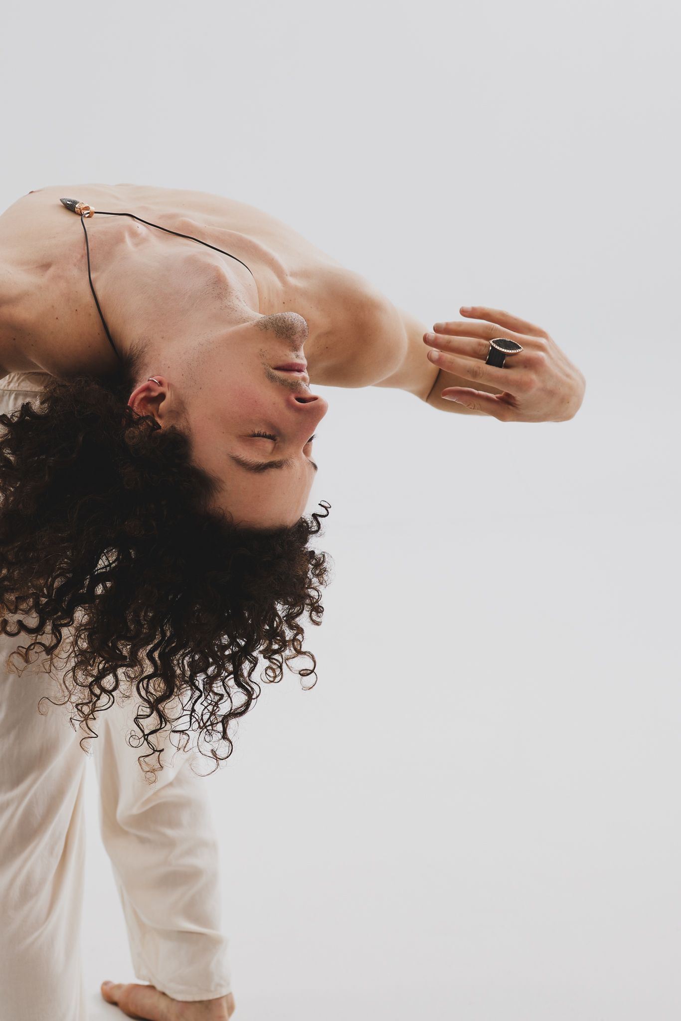 Figure bends backward gracefully with flowing curly hair, wearing white against neutral background in artistic pose.