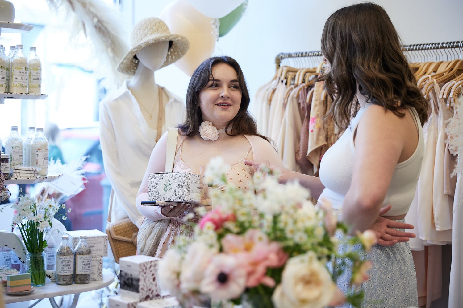 Customers browse dresses and floral arrangements in an elegant boutique with soft lighting and romantic decor.