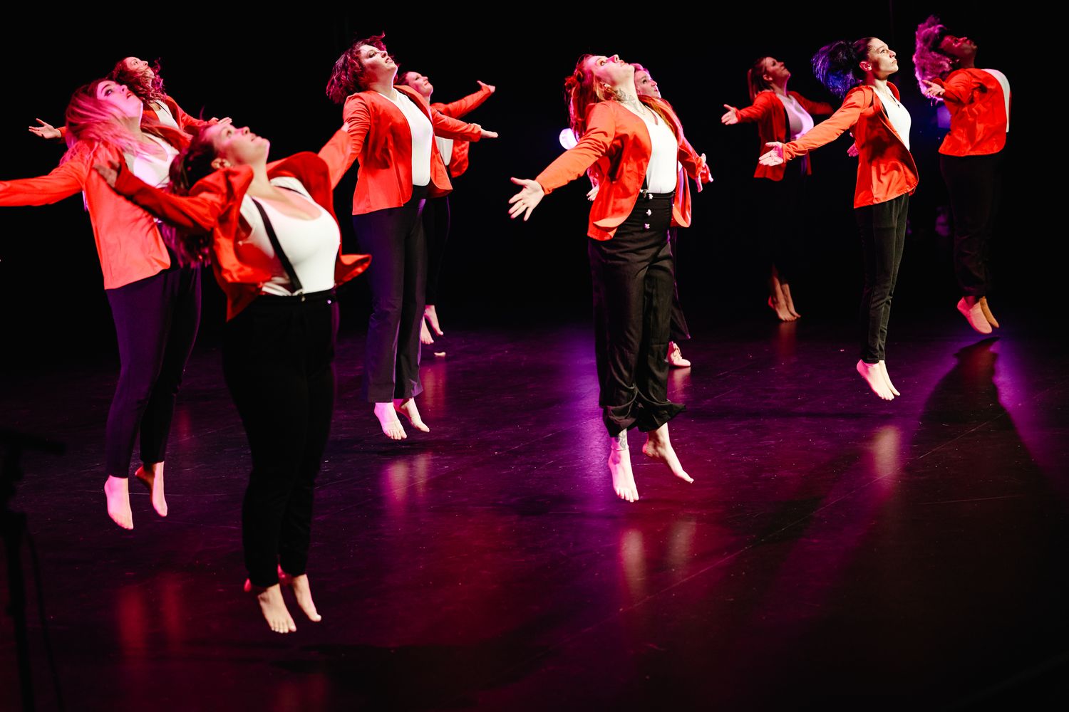 Dancers in red shirts perform a synchronized jumping routine on a dark stage illuminated by bright red lighting.