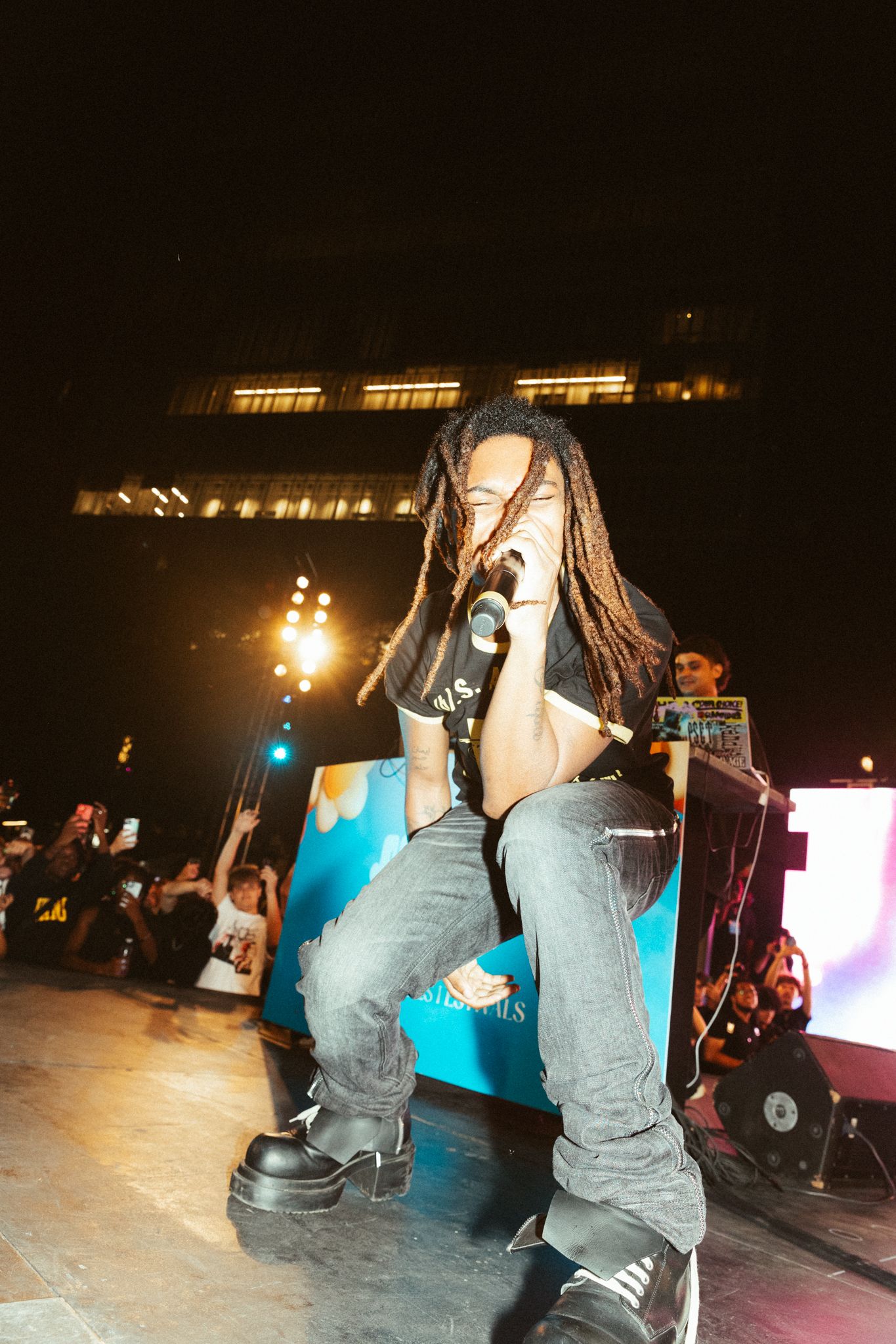 A performer with long hair sings energetically on stage under concert lighting while wearing jeans and sneakers.